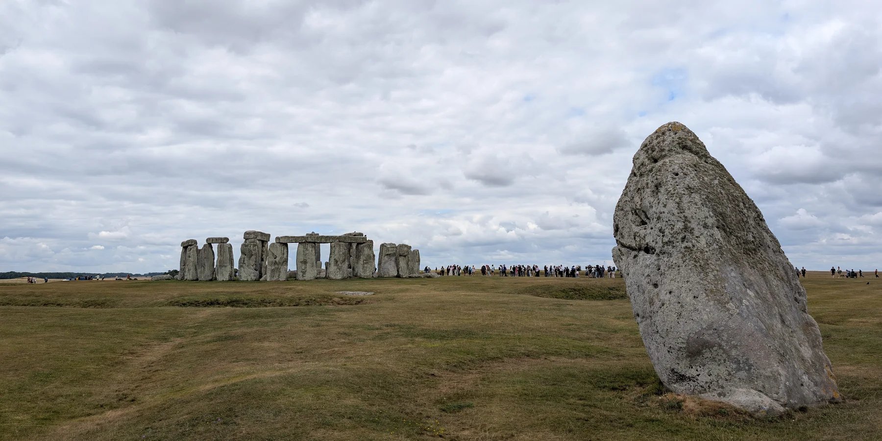 The Heel Stone in the foreground at Stonehenge, with the stone circle and a crowd of daytime visitors beyond